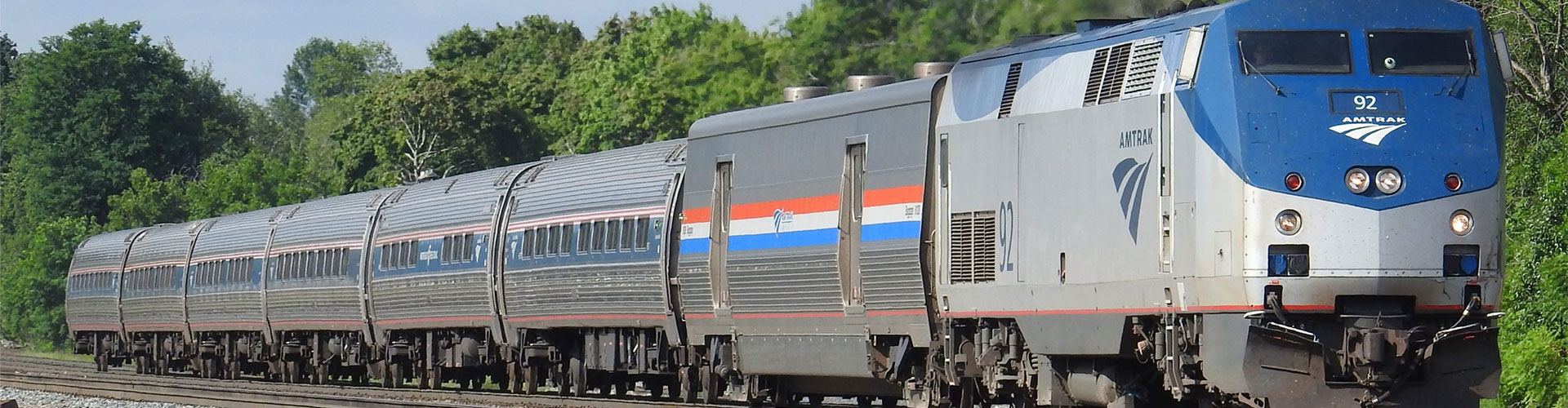 An Amtrak passenger train on the rails, with green trees in the background.