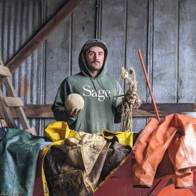 A person in a green “Sage” hoodie stands in a barn holding a cantaloupe and garlic, surrounded by farm gear on a red trailer.