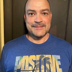 A person in a blue tee shirt standing against a wall and slightly smiling.