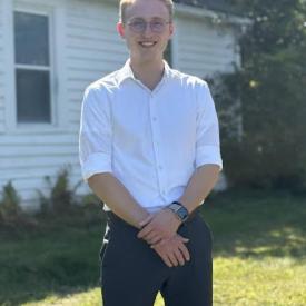 A person with short blonde hair and a white shirt stands on a lawn in front of a white building.
