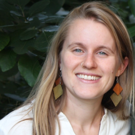 Person with long blonde hair and a white shirt smiling in front of a tree,