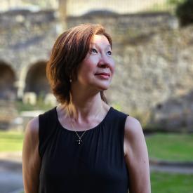 A person in a black top standing outside on a grassy lawn and looking up to the left and smiling.