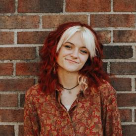 A person with red hair and a patterned shirt standing against a brick wall.