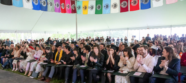 Graduates, faculty, staff members, and guests under the Commencement tent with colorful flags