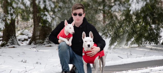 Dmitry Pepper crouching in a snowy landscape. He is holding two white dogs, both wearing red sweaters.