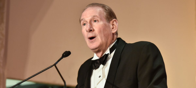 Jim Crown wears a black suit and speaks into a microphone.