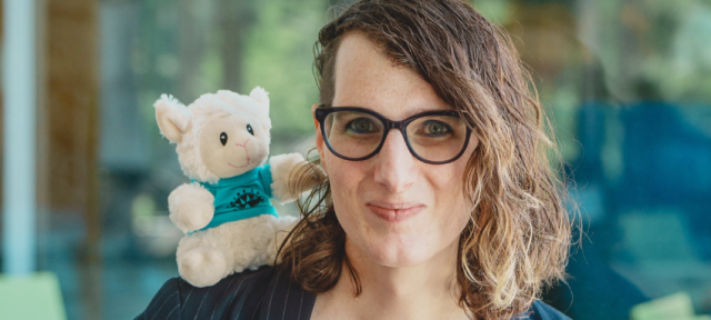 Assistant Professor of Environmental MicrobioBioremediation Juliet Johnston smiles at the camera. Sitting atop her left shoulder is a sheep stuffed animal wearing a Hampshire College tee.