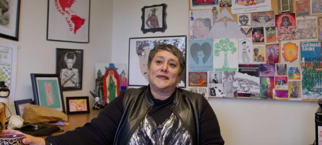 Professor of Critical Race, Gender, and Media Studies Susana Loza sits in her office. The background is full or colorful artwork.