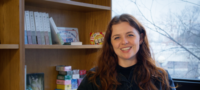 Sarah is sitting in a chair facing the camera. There is a wintery window behind her and a bookcase to her left.