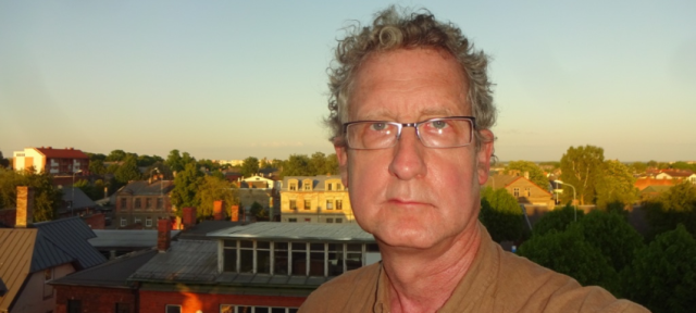 Michael Strmiska 78S is facing the camera and there is a sunset over a city in the background.