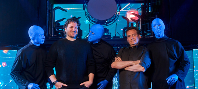 Two men stand in the center of a stage, surrounded by three performers from the Blue Man Group. The performers have their faces and hands painted blue and wear black clothing. One of them leans in toward the man on the left, who is smiling and wearing a black sweater, while the other two stand stoically. The man on the right, wearing a dark button-up shirt, crosses his arms with a slight smile. The background features a high-tech stage setup with LED screens displaying abstract visuals and circular lighting