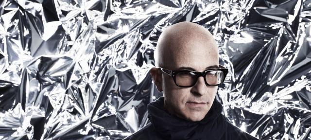 A person in glasses and a black turtleneck standing in front of a silver backdrop.