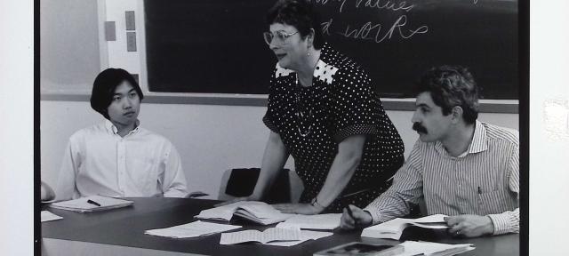 A person standing in front of a blackboard with people sitting to their right and left.