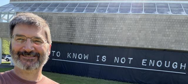 A person in glasses and a red shirt stands in front of a sign reading "to know is not enough."