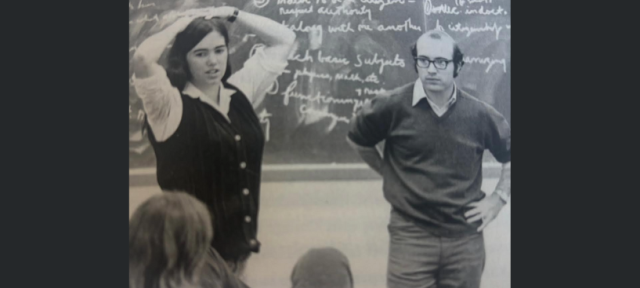 Two people stand in front of a blackboard with chalk writing on it.