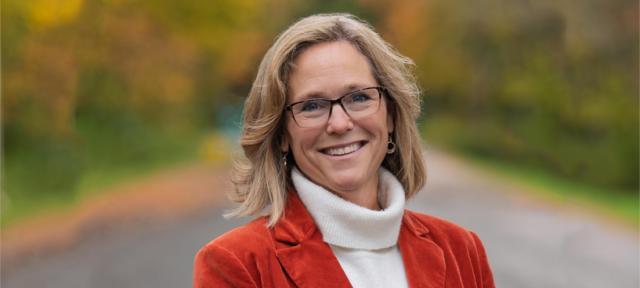 Jennifer Chrisler smiling, standing on a road with autumn foliage in the background