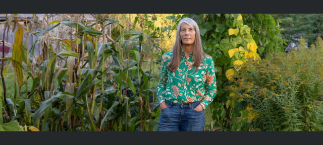 A person in a floral print shirt and jeans standing in front of a wall of plants.