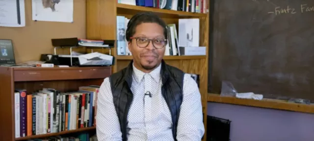 A person sitting in front of a bookshelf and chalkboard smiling.