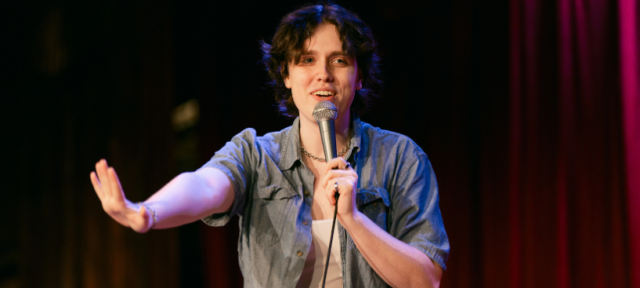 A person in a blue denim shirt over a white tank is standing on stage with a microphone in front of a red curtain smiling and holding out her hand.