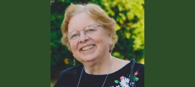 A person in glasses and a black top with flowers smiles standing outside in front of trees.