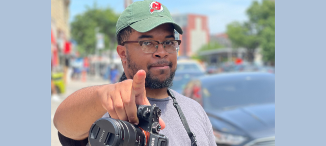 A person in a green hat and gray t-shirt pointing at the camera with another camera around their neck.