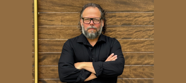 Person in glasses wearing a black button down shirt standing against a brown brick wall with arms crossed.
