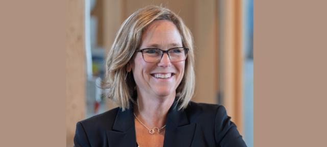 A person in a black blazer and sitting in front of an office door and smiling. 