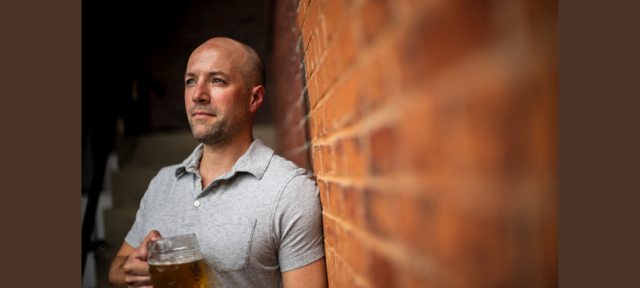 A person in a gray polo shirt standing against a brick wall holding a beer stein.