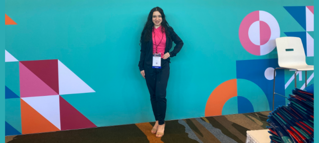 A person in a pink shirt and blue suit posing in front of a bright blue wall.