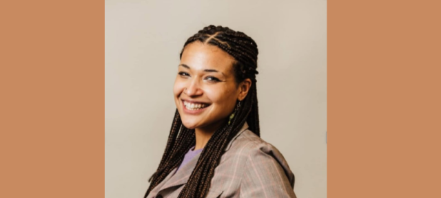A woman turning toward the camera and smiling wearing a taupe shirt in front of a beige background.
