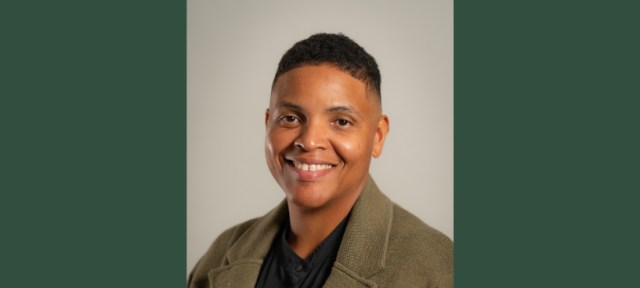 A person in a tan blazer and black shirt smiling against a plain background.