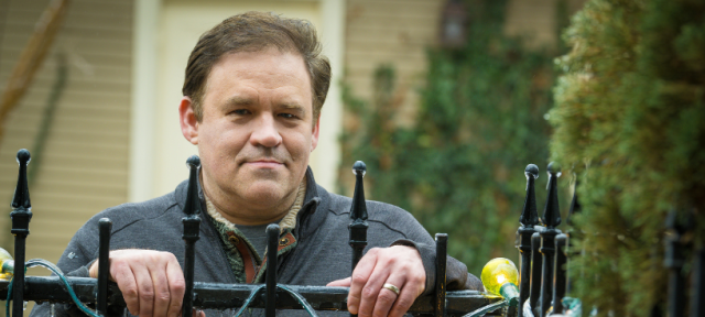 A person in a blue shirt stands behind a black iron fence and rests their hands on its top.