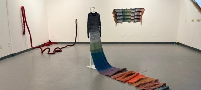An art gallery featuring a colorful knitted dress with a long train on a dress form, a long, red knitted rope hanging off the wall and onto the floor, and a  colorful knit panel on the wall.
