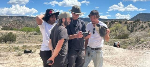 Four people standing in a huddle looking at a camera with a sweeping desert landscape behind them,