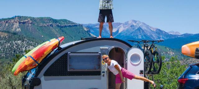 Earl stands atop camper with arms up and Glenna stands in door with leg out