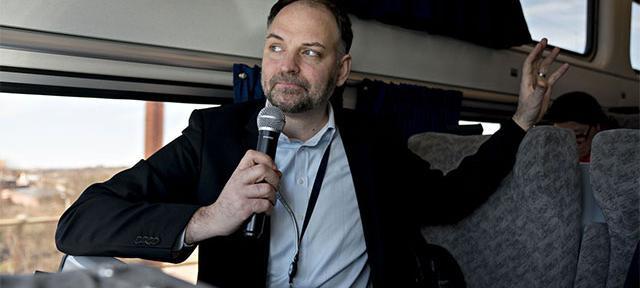 Stephen Gardner standing aboard an Amtrak train, holding a microphone in hand.