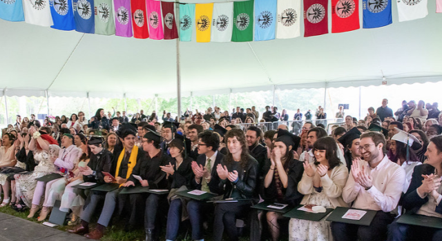 Graduates, faculty, staff members, and guests under the Commencement tent with colorful flags