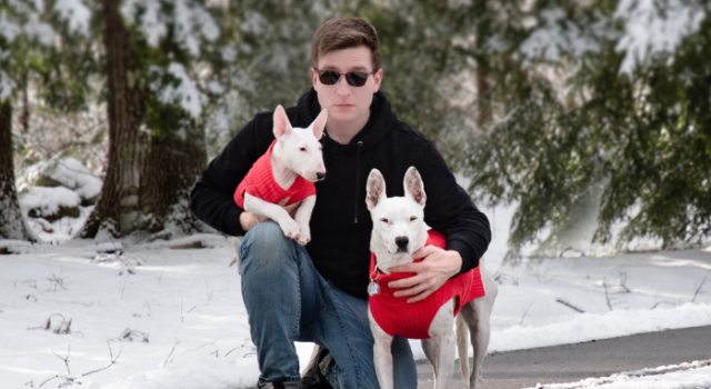 Dmitry Pepper crouching in a snowy landscape. He is holding two white dogs, both wearing red sweaters.