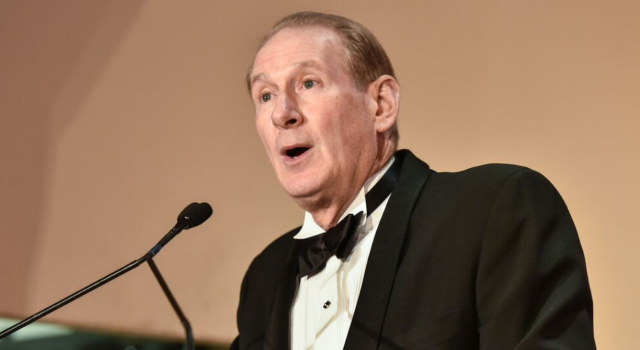 Jim Crown wears a black suit and speaks into a microphone. 