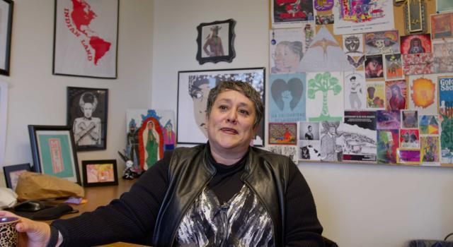 Professor of Critical Race, Gender, and Media Studies Susana Loza sits in her office. The background is full or colorful artwork.