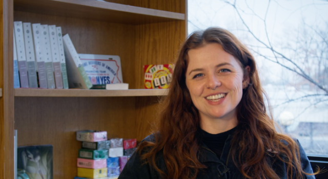 Sarah is sitting in a chair facing the camera. There is a wintery window behind her and a bookcase to her left.