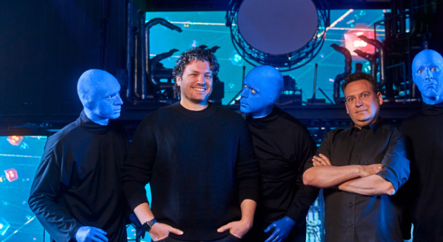 Two men stand in the center of a stage, surrounded by three performers from the Blue Man Group. The performers have their faces and hands painted blue and wear black clothing. One of them leans in toward the man on the left, who is smiling and wearing a black sweater, while the other two stand stoically. The man on the right, wearing a dark button-up shirt, crosses his arms with a slight smile. The background features a high-tech stage setup with LED screens displaying abstract visuals and circular lighting