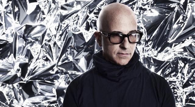 A person in glasses and a black turtleneck standing in front of a silver backdrop.