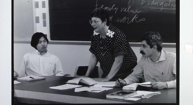 A person standing in front of a blackboard with people sitting to their right and left.