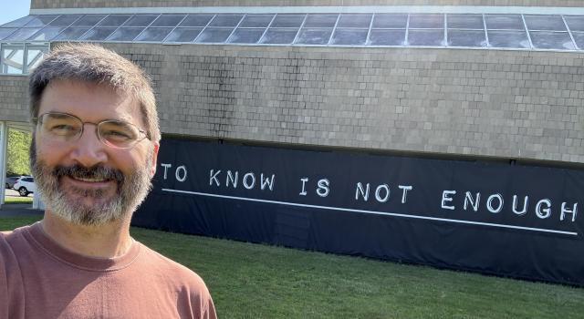 A person in glasses and a red shirt stands in front of a sign reading "to know is not enough."