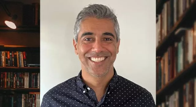 A person in a navy shirt with white dots smiling with a background of bookshelves.