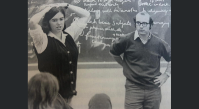 Two people stand in front of a blackboard with chalk writing on it.