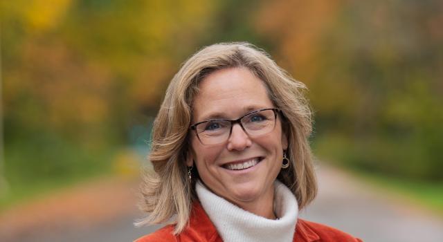 Jannifer Chrisler smiling, standing on a road with autumn foliage in the background