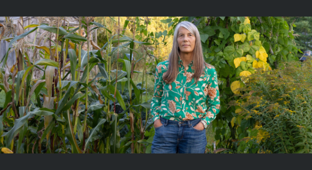 A person in a floral print shirt and jeans standing in front of a wall of plants.