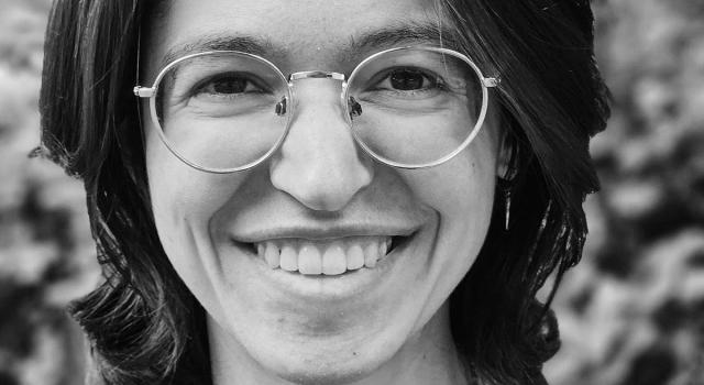 A black and white photo of a person standing in front of greenery and smiling.